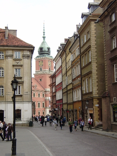 Współczesne, kolorowe zdjęcie. Przedstawia widok na ulice i kamienice, w oddali widok na wieżę zamkową. Jest to Stare Miasto w Warszawie.