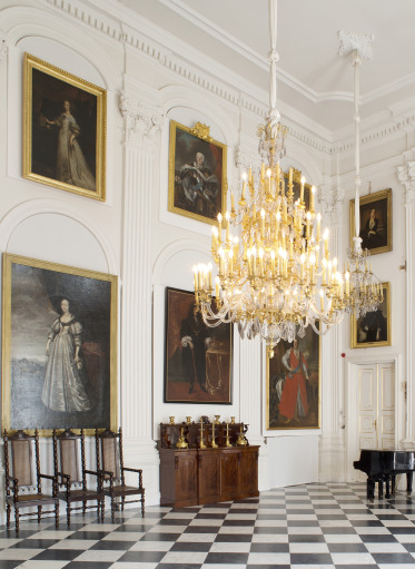 Sala pałacowa w kolorze białym ze złotym żyrandolem. Posadzka w czarno-białą szachownicę. Na ścianach wiszą obrazy w złotych ramach. Po prawej stronie stoi fortepian, po lewej stronie rząd krzeseł.