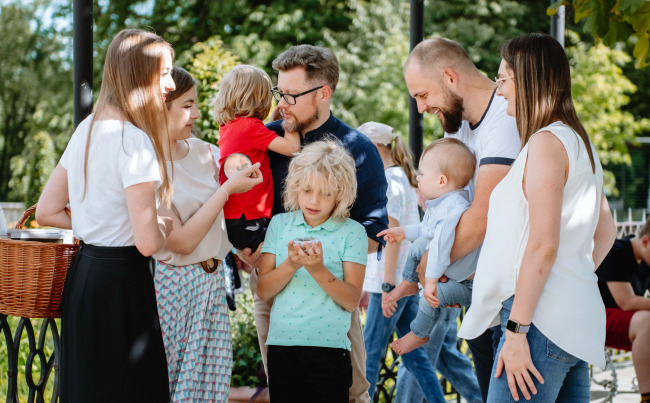 Grupa ludzi z dziećmi stoi na deptaku. Jest to spotkanie rodzin na tle pejzażu. Portretowani ubrani są w letnie stroje współczesne.